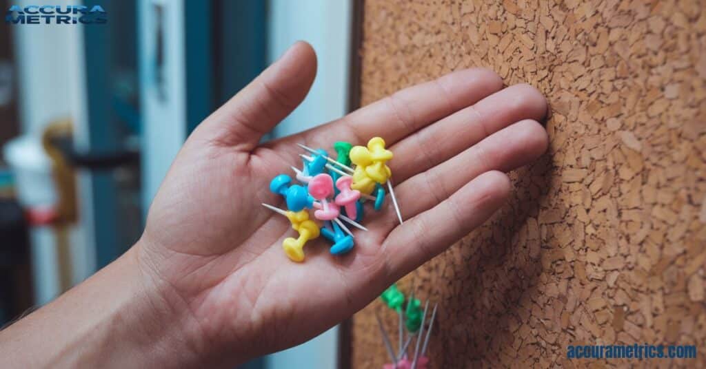 A hand holding thumbtacks near a corkboard.